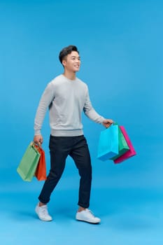 Young asian man shopaholic smiling while holding shopping bags isolated on blue