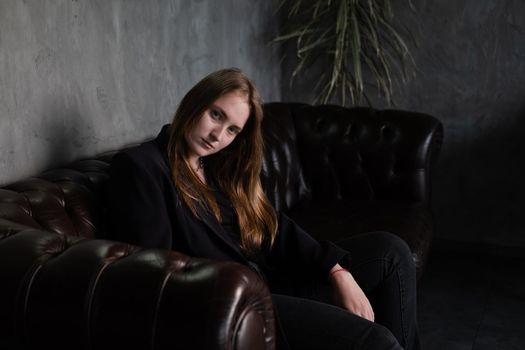 beautiful confident brunette woman in black on a brown leather couch in grey dark room. business woman. feminine. millennial people.