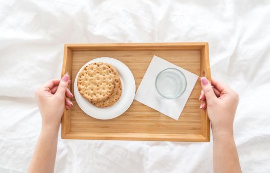 Woman holding tray with diet breakfast of crackers and water on bed