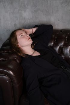 beautiful confident brunette woman in black on a brown leather couch in grey dark room. business woman. feminine. millennial people.