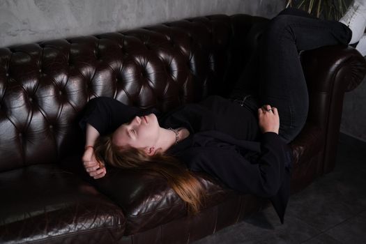 beautiful confident brunette woman in black on a brown leather couch in grey dark room. business woman. feminine. millennial people.