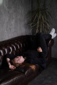 beautiful confident brunette woman in black on a brown leather couch in grey dark room. business woman. feminine. millennial people.