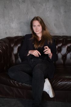 beautiful confident brunette woman in black on a brown leather couch in grey dark room. business woman. feminine. millennial people.