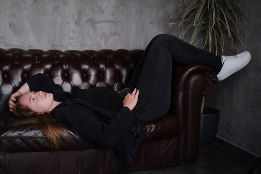 beautiful confident brunette woman in black on a brown leather couch in grey dark room. business woman. feminine. millennial people.