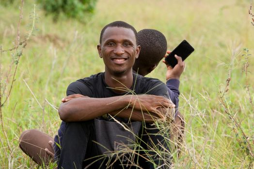 two young men leaning against each other and sitting on the grass in the park. one seen from the front smiling and the other seen from behind uses a mobile phone, Concept of friendship men.