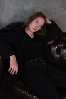 beautiful confident brunette woman in black on a brown leather couch in grey dark room. business woman. feminine. millennial people.