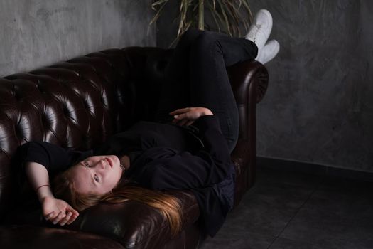 beautiful confident brunette woman in black on a brown leather couch in grey dark room. business woman. feminine. millennial people.