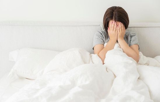 upset young brunette woman sitting on the white linen in bed at home, copyspace for your text to the left