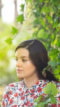 Portrait of beautiful Latin young woman looking to the left wearing floral design dress hidden among a green leafy creeper plant during the morning