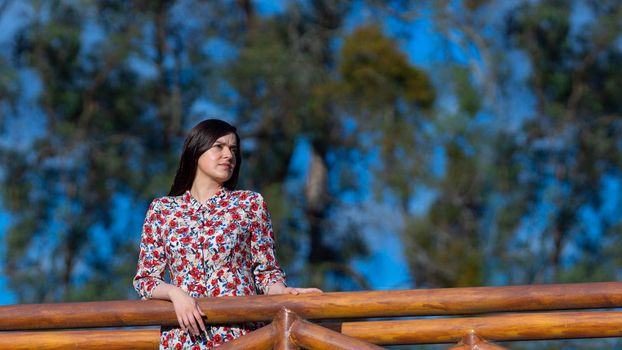 Portrait of beautiful young Latin woman looking to the right with floral design dress standing in the middle of a garden with a background of trees during the morning