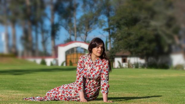 Portrait of beautiful Latin young woman looking to the right with floral design dress resting on the grass in the middle of a garden with a background of trees during the morning