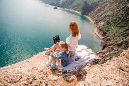 Freelance woman with her daughter working on a laptop by the sea, typing on the keyboard, enjoying the beautiful view, highlighting the idea of remote work