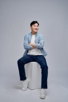 Full body young man wearing blue shirt with jeans sitting wooden cube posing in studio