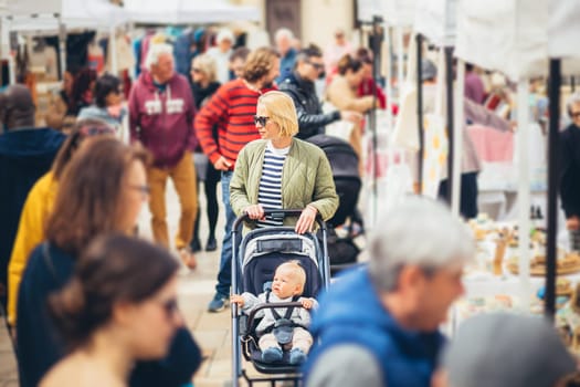 Mother waling and pushing his infant baby boy child in stroller in crowd of unrecognizable people wisiting sunday flea market in Malaga, Spain