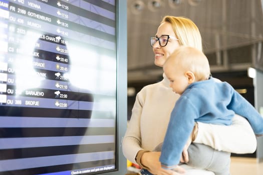 Mother traveling with child, holding his infant baby boy at airport terminal, checking flight schedule, waiting to board a plane. Travel with kids concept