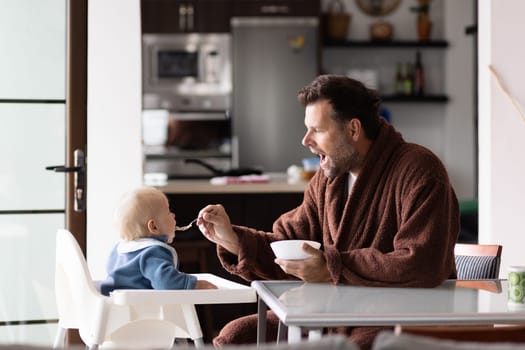 Father wearing bathrope spoon feeding hir infant baby boy child sitting in high chair at the dining table in kitchen at home in the morning.