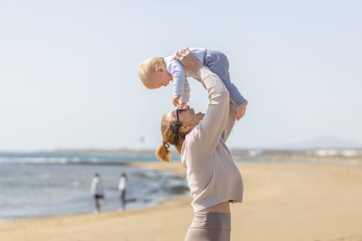 Mother enjoying summer vacations holding, playing and lifting his infant baby boy son high in the air on sandy beach on Lanzarote island, Spain. Family travel and vacations concept