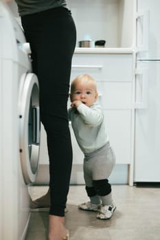 Little infant baby boy child hiding between mothers legs demanding her attention while she is multitasking, trying to do some household chores in kitchen at home. Mother on maternity leave