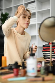 Shot of young gay man applying moisturizing serum product on face, doing daily makeup in living room. LGBTQ, lifestyle concept.