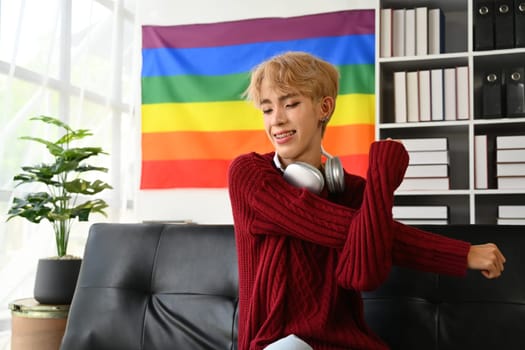 Happy young gay man relaxing on couch at home with closed eyes, stretching arms. LGBTQ, people lifestyle, leisure concept.