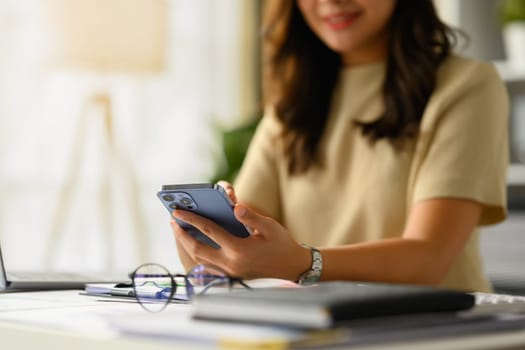 Smiling young woman sitting at working desk, texting messages, chatting in social on smartphone. Select focus on hands.