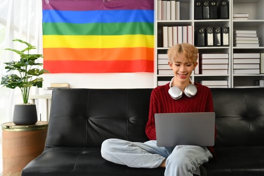 Delighted young gay man using laptop on couch in living room with LGBT pride rainbow flag on background.