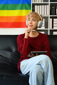 Happy teenage man wearing wireless headphone using digital tablet in bright living room with rainbow pride flag.