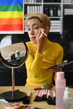 Beautiful gay man doing daily makeup in living room, applying eye shadow with brush. LGBTQ, lifestyle and makeup concept.