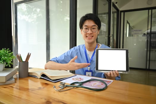 Young surgeon wearing blue scrubs holding and showing digital tablet with blank screen. Healthcare and medical concept.