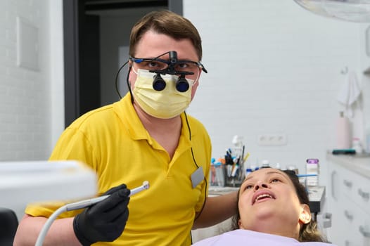 Professional dentist wearing protective medical mask and orthodontist binoculars, holds dental drill and looks at camera while performs teeth treatment to pregnant female patient in dentistry clinic