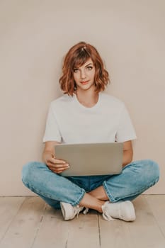 A brunette sits on the floor with a laptop on a beige wall background. She is wearing a white T-shirt, jeans and white sneakers