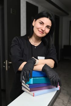 Confident beautiful woman, experienced professional doctor dentist, smiling looking at camera, standing at reception counter with a stack of medical books and encyclopedia in a modern dentistry clinic