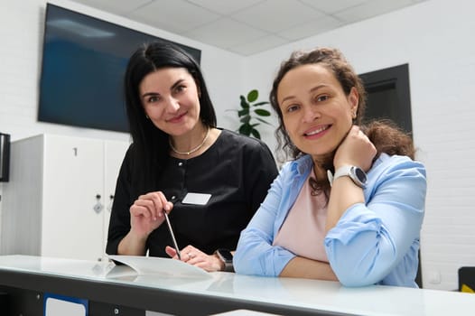 Beautiful woman, confident dentist doctor, dental hygienist or female orthodontist and her happy pregnant patient ,smiling looking at camera, standing at reception counter in modern dental clinic