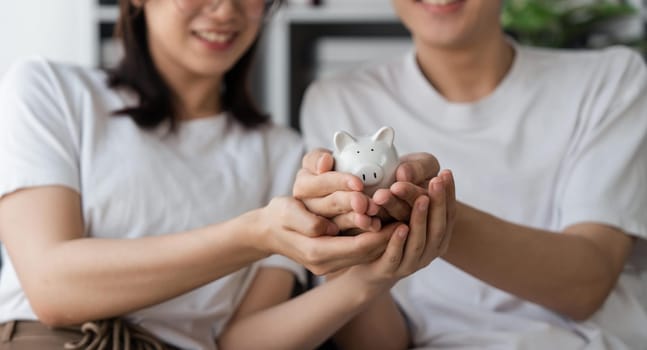 Happy asian young couple love calculate and putting coin in piggy bank for saving money to buy real estate for new home. Business finance, deposit with banking for financial planning future together.