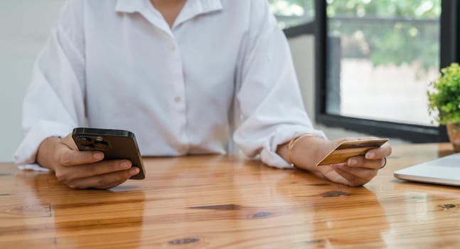 Young adult Asian female consumer holding credit card and smartphone at home doing online banking transaction. E commerce virtual shopping, secure mobile banking concept.