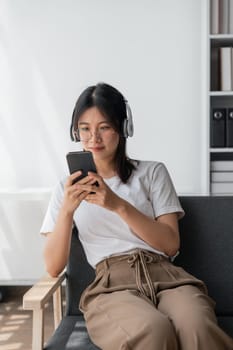 Young Asians woman listening to music on couch in living room at home. Happy Asia female using mobile smartphone, wearing headset and sitting on sofa.