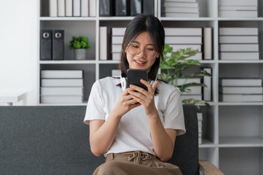 Happy Asian beautiful woman is using mobile phone sitting on a sofa at home.