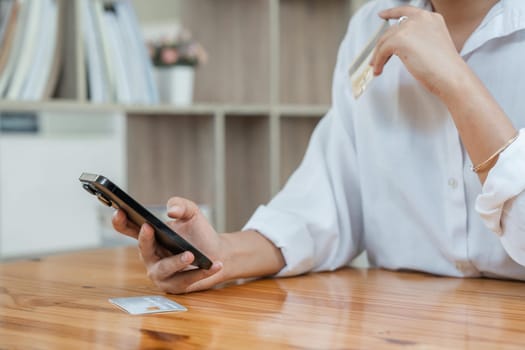 Young adult Asian female consumer holding credit card and smartphone at home doing online banking transaction. E commerce virtual shopping, secure mobile banking concept.