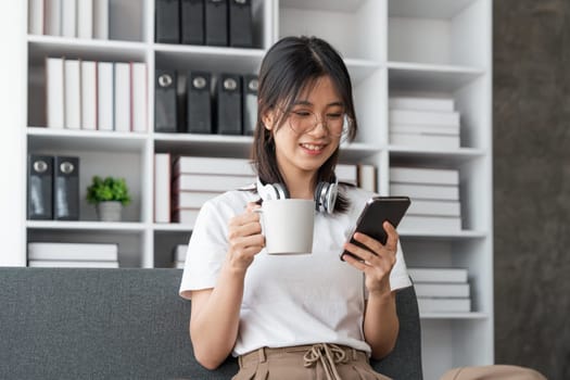 Happy Asian beautiful woman is using mobile phone sitting on a sofa at home.