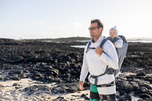 Young father carrying his infant baby boy son in backpack on black rock volcanic beach on Lanzarote island, Spain. Family travel and winter vacation concept