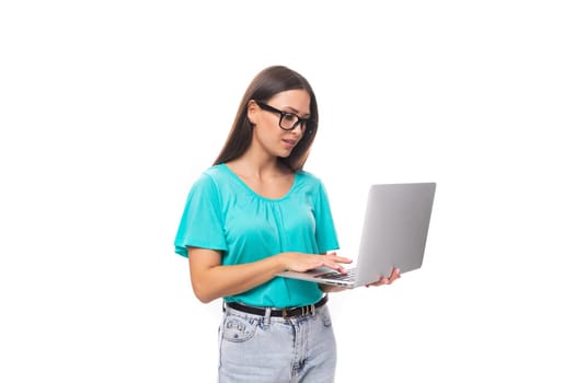 young smart caucasian female model in glasses with black silky smooth hair dressed in a blue t-shirt works using the internet and a laptop.