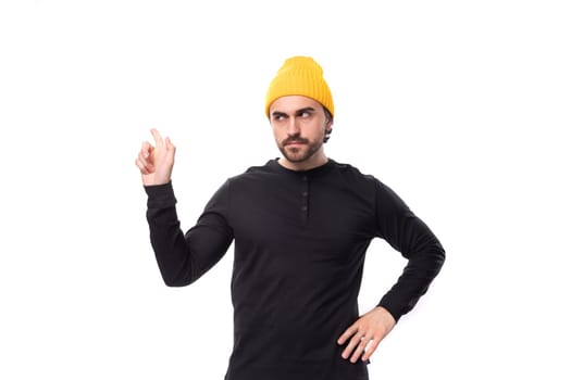 a brutal handsome young 30 year old guy dressed in a black jacket and a yellow hat points to the wall with his hand.