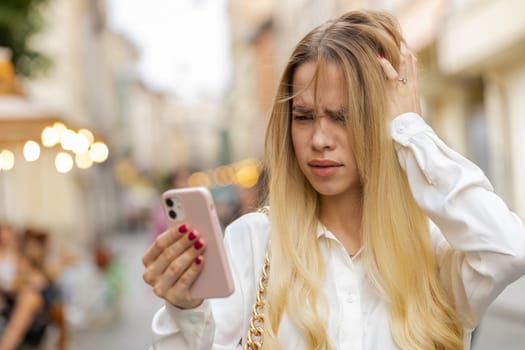 Blonde young woman use smartphone typing browsing, loses becoming surprised sudden lottery results, bad news, fortune loss, fail. Teenager girl walking in urban city street outdoors. Town lifestyles