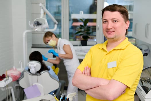 Confident portrait of a Caucasian young happy male dentist orthodontist in yellow medical uniform, standing with arms folded in the dental office in modern dentistry clinic, smiling looking at camera