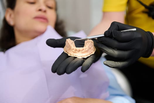 Closeup gloved hands of orthodontist holding dental plaster mold over blurred background of a woman in dentist's chair. Dentist showing at camera the patient's dental gypsum models. Dentistry. Surgery
