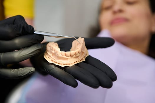 Dentist showing dental plaster mold to the camera, holding hands above a female patient lying in dentist's chair, getting treatment in dentistry clinic. Jaw model. Health, dental and oral care concept