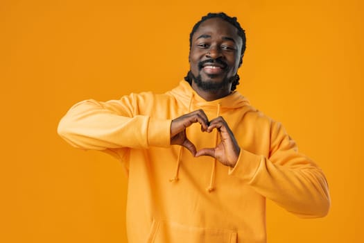 Young african man with beard wearing casual sweatshirt doing heart symbol shape with hands on yellow