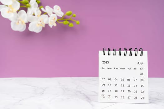 July 2023 desk calendar and white orchid flower on wooden background.