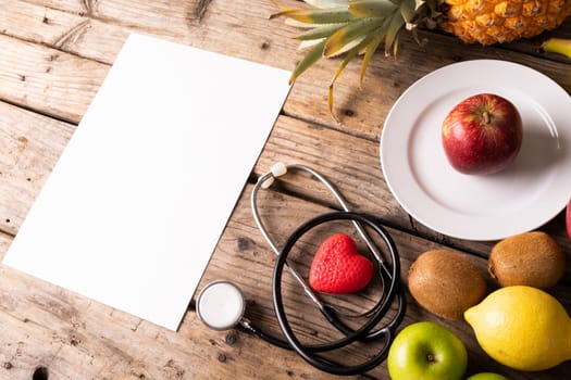 High angle view of various fruits with stethoscope and heart shape by blank paper on wooden table. unaltered, copy space, healthy food, variation and organic concept.