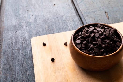 High angle view of chocolate chips in bowl on cutting board over wooden table. unaltered, sweet food and unhealthy eating concept.